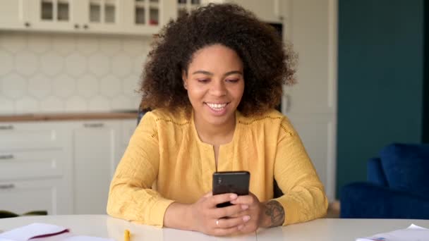 Безмятежная афроамериканка сидит за столом дома и пользуется смартфоном — стоковое видео
