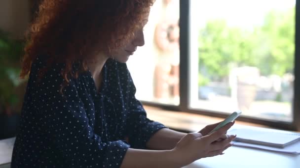 Kaukasische vrouw met krullend kapsel zitten aan het bureau en met behulp van smartphone — Stockvideo