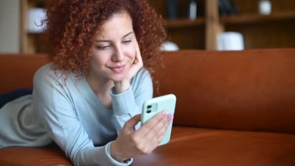 可愛いです赤毛女性使用して携帯電話下に横たわっザ居心地の良いソファ — ストック動画
