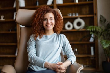 Çekici kızıl saçlı kadın koltukta oturuyor ve kameraya bakıyor.