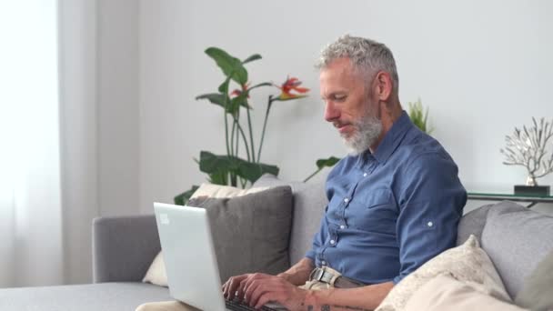 Hipster mittleren Alters im schicken lässigen Hemd mit Laptop auf der heimischen Couch — Stockvideo