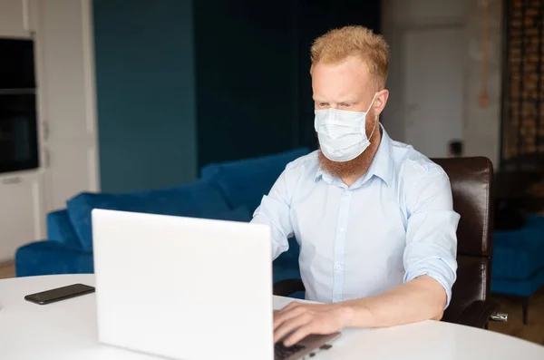 Concentrated redhead bearded man wearing protective medical mask using laptop — 图库照片