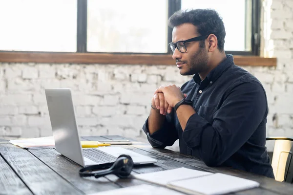 Pensativo hindú freelancer tipo con gafas mirando a la pantalla del ordenador portátil —  Fotos de Stock