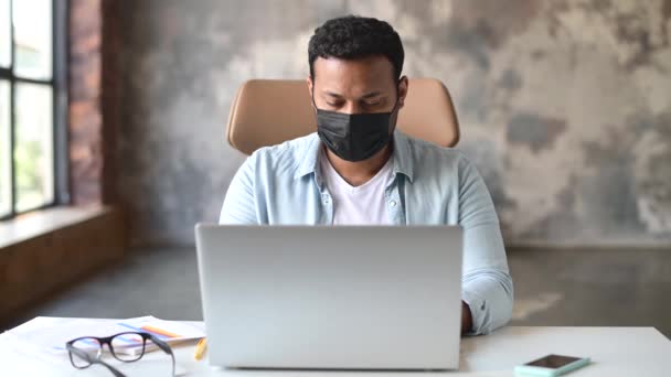 Confident indian young freelancer man sits at the desk wearing protective mask — Vídeos de Stock