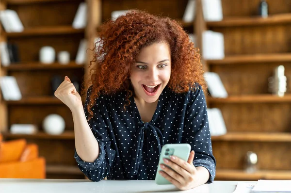 Удивлённая кудрявая рыжая молодая женщина, пользующаяся смартфоном в помещении — стоковое фото