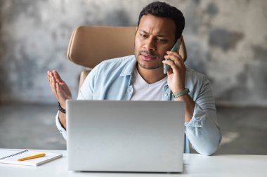 Sinirli Hintli adam akıllı telefonda konuşuyor. Masada dizüstü bilgisayarla oturuyor.