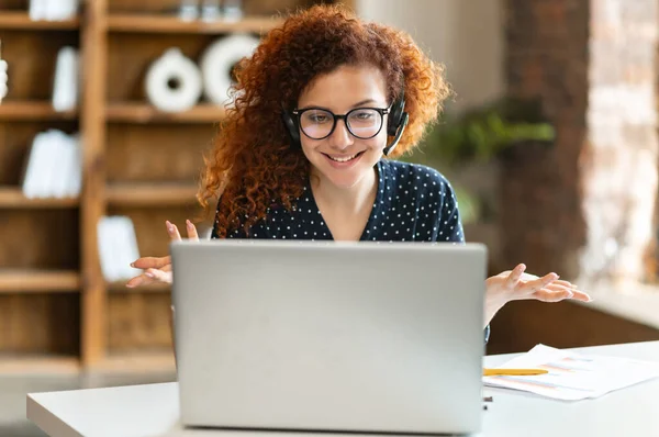 Sonriente de pelo rojo rizado mujer con auriculares mira a la webcam de la computadora portátil y hablando en línea — Foto de Stock