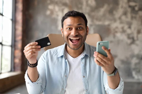 Homem indiano chocado segurando cartão de crédito e smartphone, olha para a câmera — Fotografia de Stock