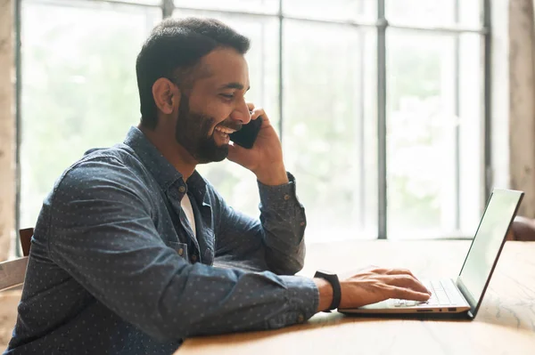 Ragazzo freelance indiano ottimista e allegro che parla su smartphone e utilizza il computer portatile — Foto Stock