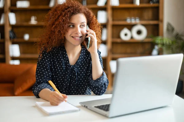 Mulher usando o laptop, falando no smartphone e fazendo anotações — Fotografia de Stock