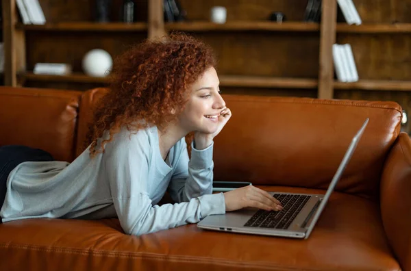 Donna trascorre il tempo libero online, donna sorridente sdraiata sul divano con un computer portatile — Foto Stock