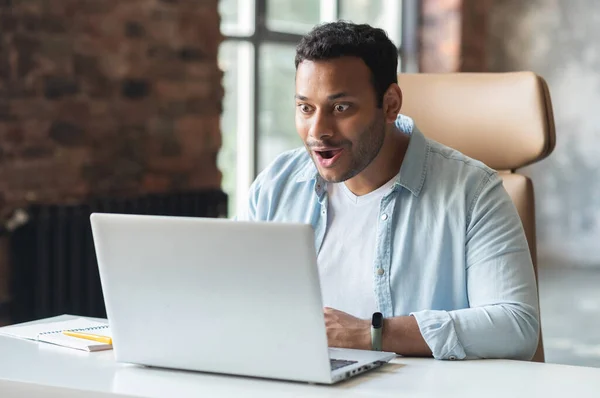 Excited surprised indian dark-haired man in casual shirt staring at laptop