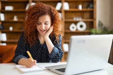 Gülümseyen, kıvırcık, kızıl saçlı bir kadın akıllı telefonda dizüstü bilgisayar kullanarak konuşuyor.