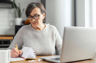 Güzel bir emekli kadın mutfaktaki masada dizüstü bilgisayar kullanıyor.
