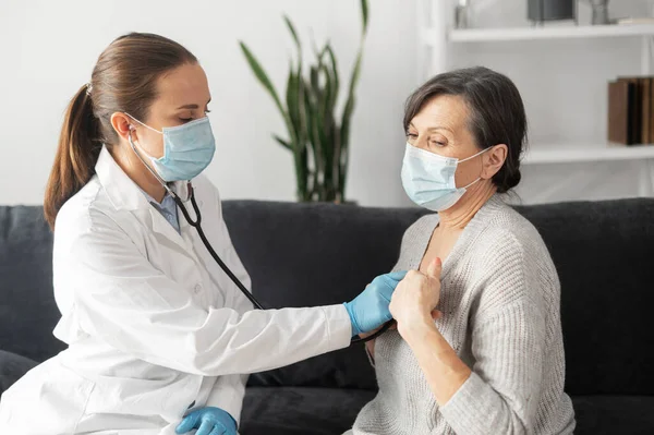 Terapeuta em máscara médica protetora e vestido médico visita seu paciente sênior em casa — Fotografia de Stock