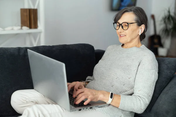 Чарівна сучасна старша жінка використовує ноутбук, сидячи на зручному дивані — стокове фото