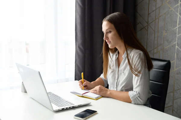 Uma jovem mulher sorrindo no desgaste casual senta-se na mesa na frente do laptop na moda — Fotografia de Stock