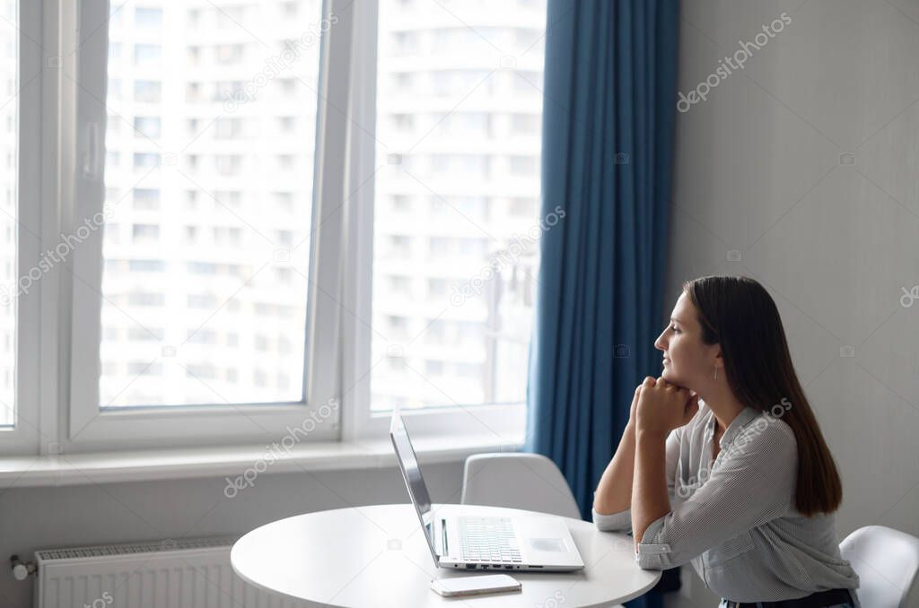 Carefree and tranquil young woman sitting at the desk and using laptop computer looks away