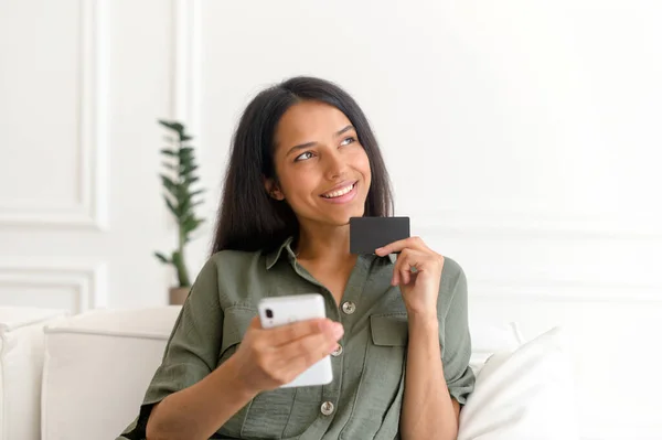 Serene sorrindo sonhador mulher indiana segurando cartão de crédito e smartphone — Fotografia de Stock