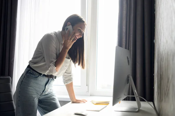 Masanın yanında duran, akıllı telefonla konuşan ve bilgisayar ekranına bakan bir kadın. — Stok fotoğraf
