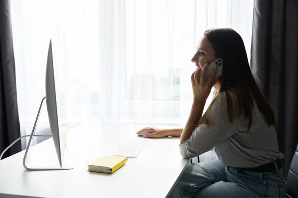 Joven freelancer mujer está hablando en el teléfono inteligente trabajando con el ordenador portátil de la oficina en casa —  Fotos de Stock