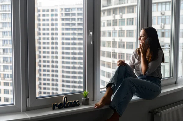 Charming woman talking on the smartphone sitting on the window sill at home