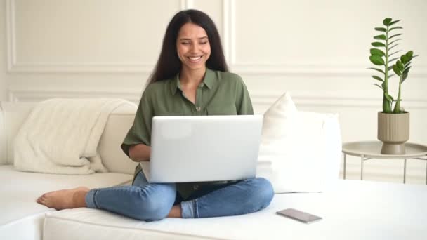 Sonriente optimista joven india freelancer o una estudiante sentada en el sofá con portátil — Vídeo de stock