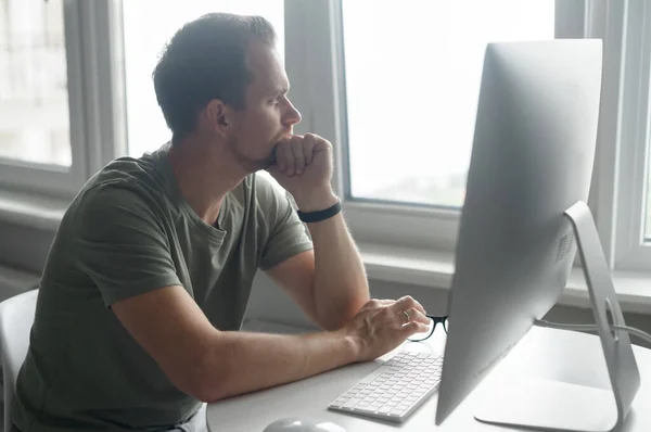 Pencive intelligente jongeman in casual t-shirt zit aan het bureau met trendy PC — Stockfoto