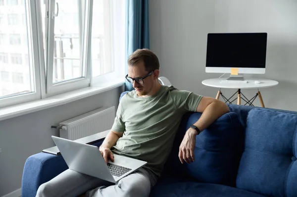 Красивый молодой европейский мужчина с бородой сидит на диване с ноутбуком — стоковое фото