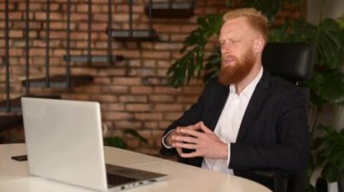 Resmi takım elbiseli, kızıl saçlı, sakallı bir iş adamı laptopta video görüşmesi yapıyor.