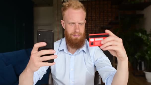 Ruiva barbudo homem em camisa casual inteligente segurando cartão de crédito e smartphone — Vídeo de Stock