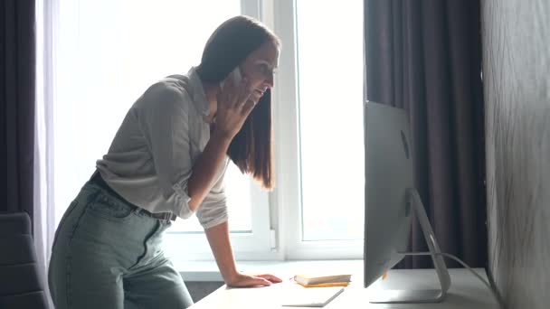 Sorridente giovane donna freelance, dipendente è in piedi vicino al posto di lavoro, parlando al telefono — Video Stock