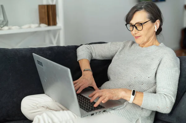 Обаятельная современная пожилая женщина с ноутбуком, сидящая на удобном диване — стоковое фото