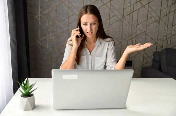 Mujer irritada hablando por teléfono mientras está sentada en el escritorio de la oficina en casa —  Fotos de Stock