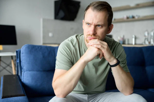 Worried guy sits on the sofa alone and looks away lost in thoughts — Stock Photo, Image