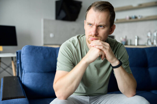 Worried guy sits on the sofa alone and looks away lost in thoughts