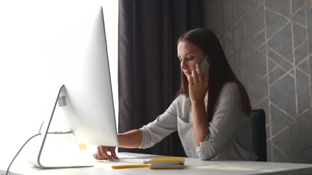 Jovem freelancer mulher está falando no smartphone trabalhando com laptop de casa escritório — Vídeo de Stock