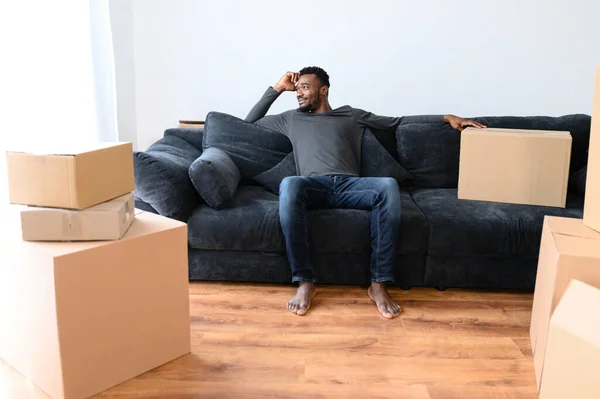 Satisfied African guy sitting on the comfortable couch among boxes — Stock Photo, Image