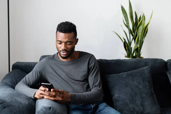 Афроамериканец сидит на диване и пользуется смартфоном. — стоковое фото