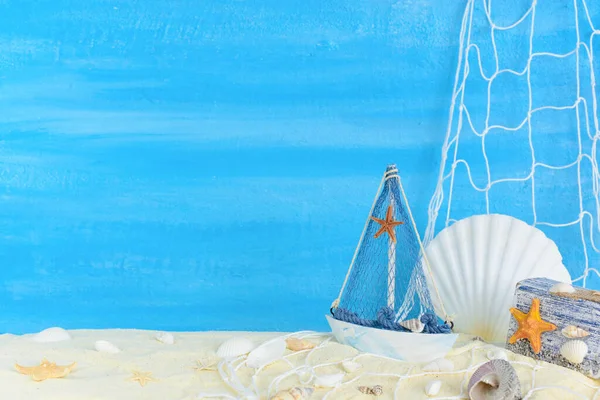 Ship, fishing net, seashells, starfish and chest on sand with copy space on blue textured backdrop.