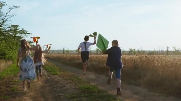 Glückliche Kindheit, Freunde werfen Papierflieger in den Himmel, während sie auf einer Landstraße in der Nähe des goldenen Weizenfeldes auf dem Land laufen — Stockvideo