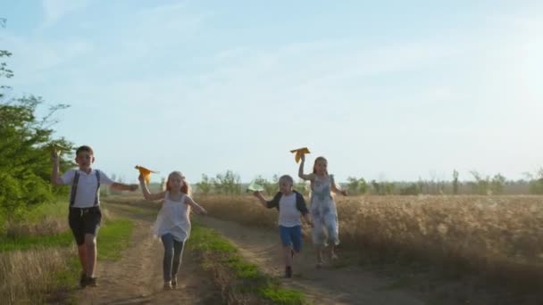 Gruppe von Freunden, Jungen und Mädchen, die entlang einer staubigen Landstraße laufen und Papierflieger in die Luft werfen, in der Nähe des goldenen Weizenfeldes — Stockvideo