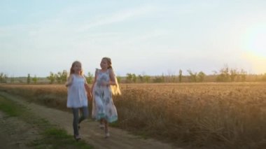 Neşeli bir tatil, uzun sarı saçlı mutlu kızlar gün batımında buğday tarlasında el ele yürürken zıplıyorlar.