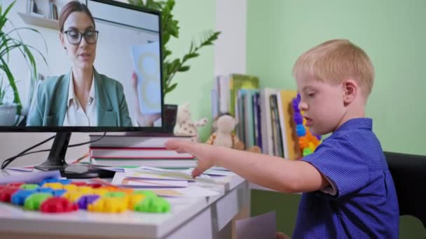 Online utbildning, pojke med ner syndrom visa kort under lärande skola läxor till kvinnliga lärare hemifrån vid bordet med en dator — Stockvideo