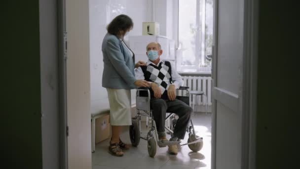 बुजुर्ग पत्नी अस्पताल में व्हीलचेयर पर बैठे वरिष्ठ पति पर हाथ डाल रही है, चिकित्सा मास्क में पुराने विवाहित दादा-दादा सेवानिवृत्ति में एक दूसरे का समर्थन करते हैं — स्टॉक वीडियो