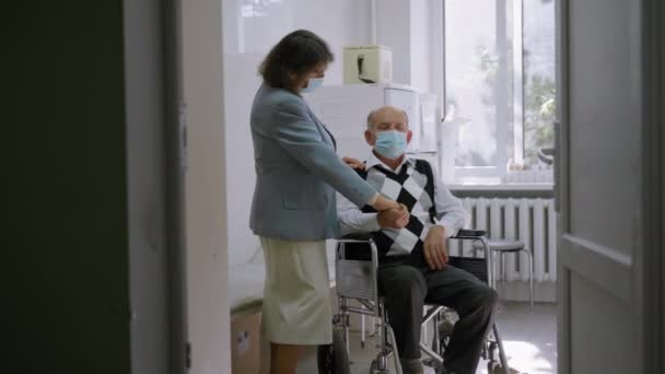 Älterer grauhaariger Mann im Rollstuhl unterhält sich mit reifer Frau, die ihn bei Krankheit im Krankenhaus unterstützt, älteres Paar in medizinischen Schutzmasken hält sich die Hände — Stockvideo