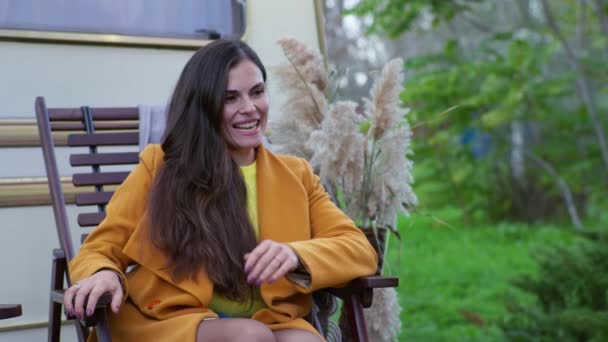 Усміхнена жінка весело сміється, сидячи на дерев'яному стільці біля кемпера і з любов'ю дивиться на свою сім'ю, осінні канікули на відкритому повітрі — стокове відео