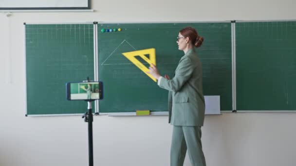 在线培训，戴眼镜的老师，手里拿着尺子，在学校靠近黑板的地方教数学，还用相机手机说话 — 图库视频影像