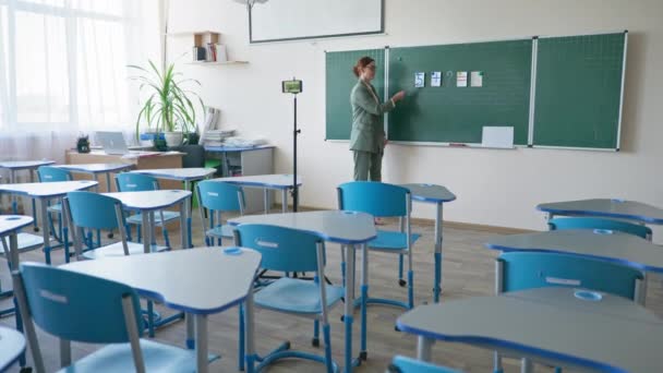 Lehrerin in leerem Klassenzimmer in der Nähe der Tafel mit Handy-Videokamera, die sich während des Online-Unterrichts aufzeichnet — Stockvideo