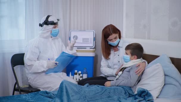 Virüs ve enfeksiyondan korunmak için koruyucu maske ve giysi takan kadın tıp çalışanı, temassız termometreyle düşük ısıyı ölçen sağlıksız bir çocuğu muayene ediyor. — Stok video